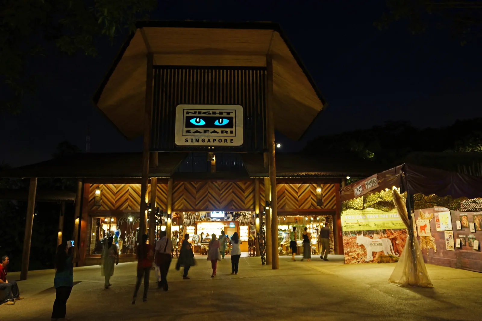 The lit entrance pavilion of the Night Safari at night, with a pitched wooden roof, a blue sign above the doors and visitors walking in